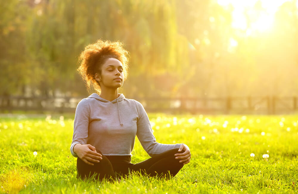 Mulher negra meditando na grama embaixo da luz do sol.