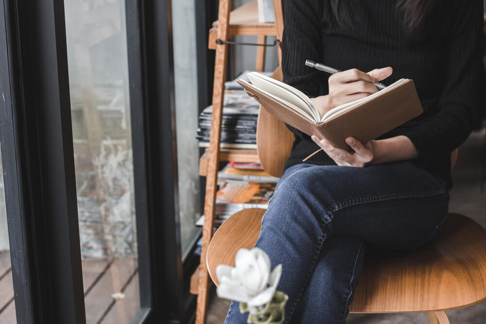Pessoa sentada em uma cadeira e escrevendo à mão em um caderno.