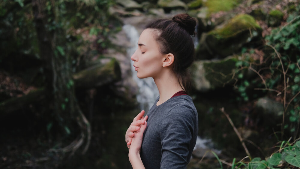 Mulher em pé, com as mãos no peito, meditando na beira de uma cachoeira.