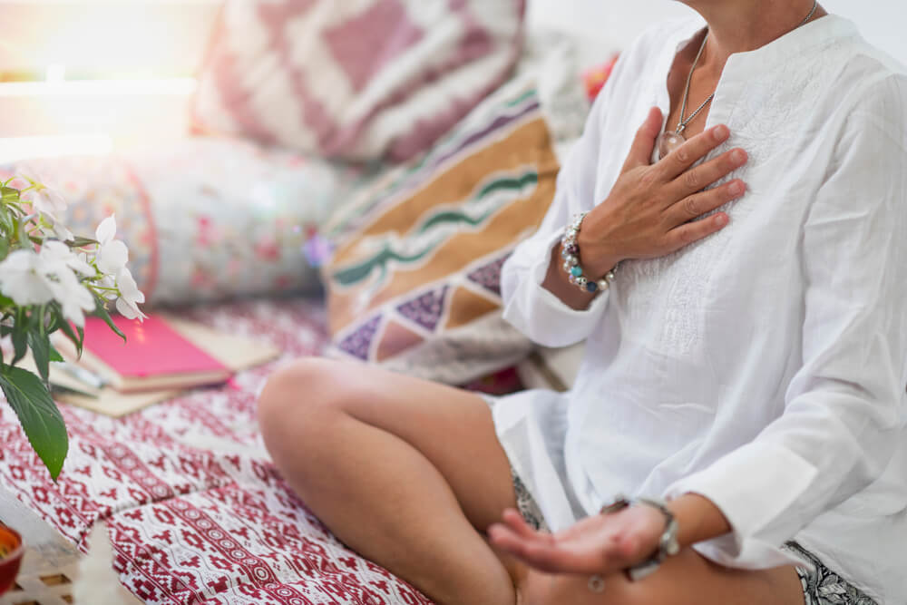 Mulher com bata branca e colar de pedra meditando com a mão no peito.