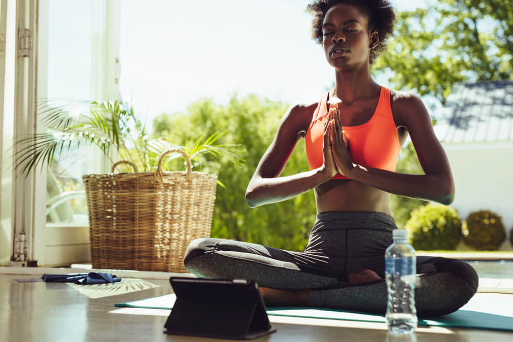 Mulher negra sentada de pernas cruzadas e com as mãos unidas, meditando.