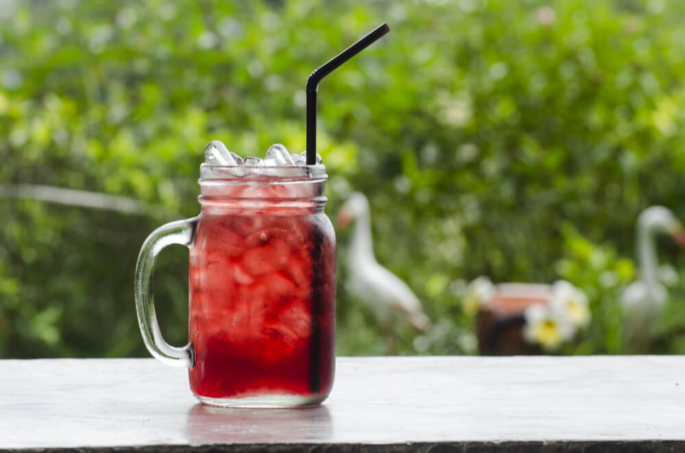 Caneca cheia de gelo e de chá de hibisco gelado, com canudo preto, sobre a mesa.